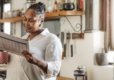 Smiling African American woman drinking a cup of coffee and reading the newspaper while standing in her kitchen in the morning